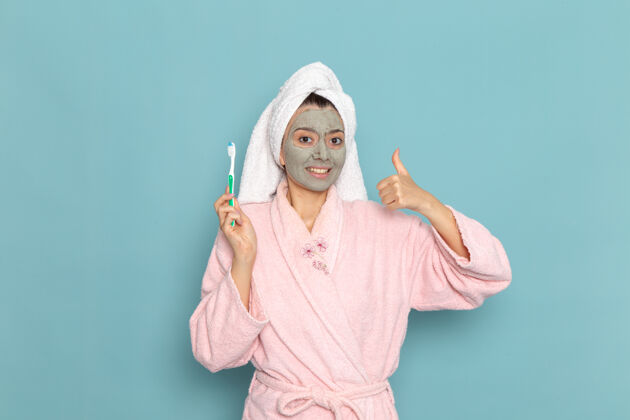 视图正面图身着粉色浴袍的年轻女性手持牙刷在蓝色墙壁上清洁美丽干净的水自我护理霜淋浴牙刷女性清洁