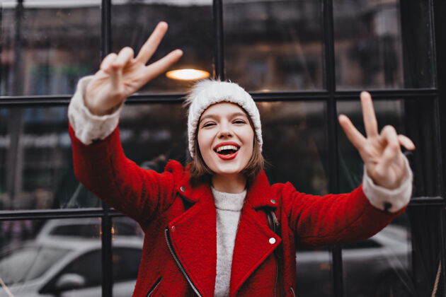 肖像戴着针织帽子 穿着红色保暖夹克的女人微笑着 展示着和平的标志 在黑色木框窗户的背景下看着镜头外套年轻天气