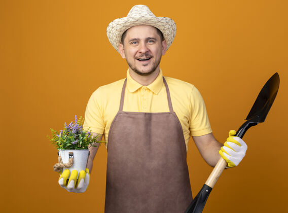 成人年轻的园丁穿着连体衣 戴着帽子 拿着铲子和盆栽植物 站在橙色的墙上 面带微笑地看着前方微笑工作服花