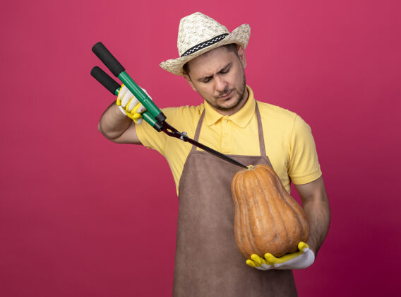 手套年轻的园丁穿着连体衣 戴着帽子 戴着工作手套 手里拿着南瓜 站在粉红色的墙上用树篱剪剪南瓜帽子制服人