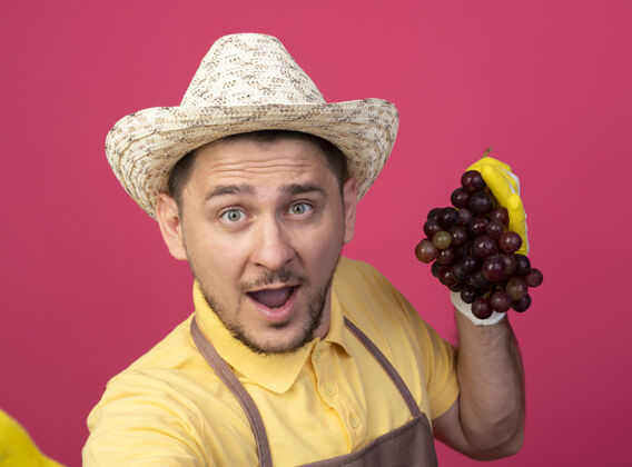 抱年轻的园丁穿着连体衣 戴着帽子 戴着工作手套 手里拿着一束葡萄 站在粉色的墙上惊讶地看着前方束人男