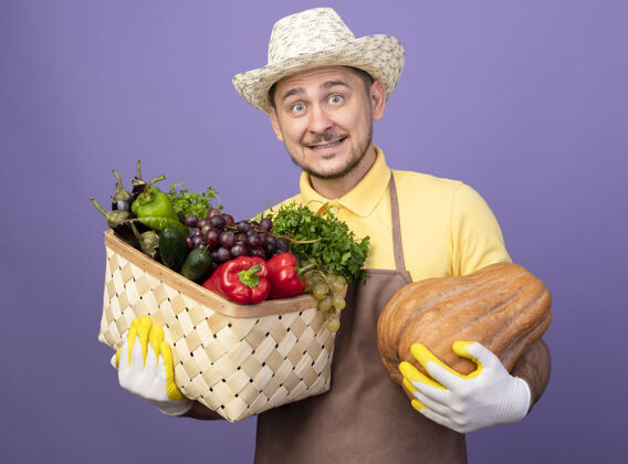 帽子年轻的园丁穿着连体衣 戴着帽子 戴着工作手套 手里拿着装满蔬菜的箱子 手里拿着南瓜 面带微笑地看着前面 站在紫色的墙上 满脸幸福穿工作年轻