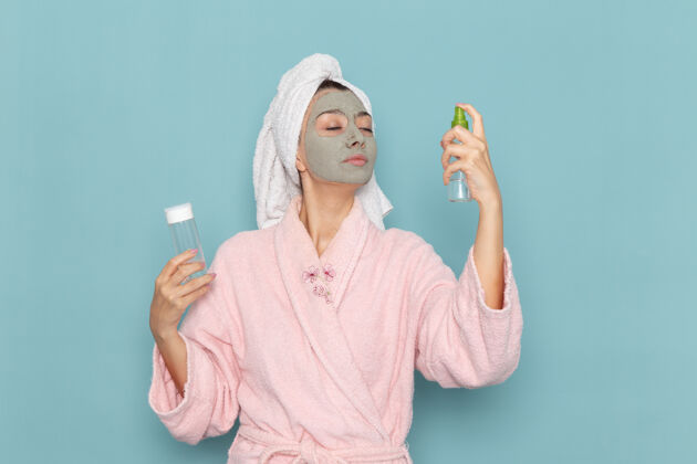 女性正面图身着粉色浴袍的年轻女性淋浴后拿着喷在蓝色墙上的美丽水浴霜自理淋浴淋浴人浴袍