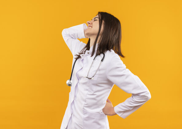 公民年轻的女医生穿着白大褂 脖子上戴着听诊器 看起来不舒服 摸着背 站在橘色的墙上感到疼痛工作人听诊器