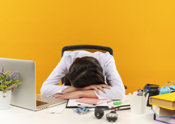 女性身穿白大褂的年轻女医生躺在桌子上 橙色的墙上挂着笔记本电脑和文件人人年轻人