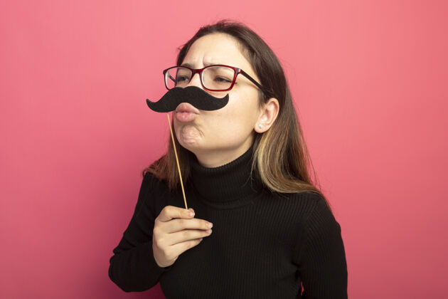 吹一个穿着黑色高领毛衣戴着眼镜的快乐的年轻漂亮女人 手里拿着一把有趣的小胡子 站在粉色的墙上吹着一个吻漂亮搞笑胡子