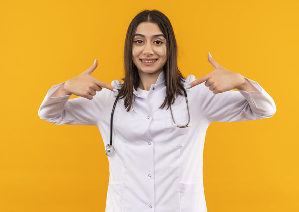 外套年轻的女医生穿着白大褂 脖子上戴着听诊器 用食指指着站在橙色墙上微笑的自己医生姿势脖子