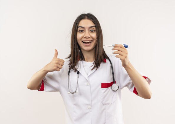 外套年轻的女医生穿着白大褂 脖子上戴着听诊器 手持数字体温计 微笑着竖起大拇指站在白墙上表演人听诊器