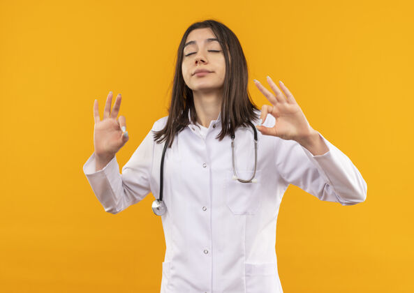 医生年轻的女医生穿着白大褂 脖子上戴着听诊器 闭着眼睛放松 做着冥想的手势 手指站在橙色的墙上闭上外套脖子