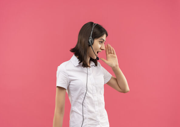 有人穿着白衬衫 戴着耳机的年轻女孩 站在粉红色的墙上 用手在嘴边喊着或叫着什么人人手耳机