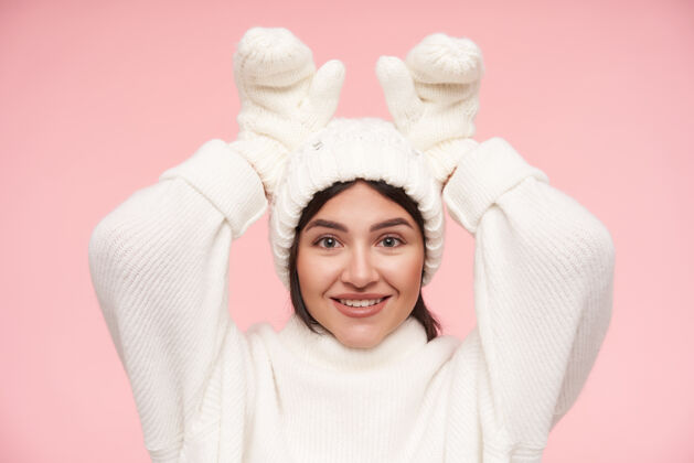年轻快乐的年轻迷人的棕色头发的女人穿着白色毛衣 戴着手套和帽子 在粉红色的墙上摆姿势 举起手来模仿发髻的耳朵发型兔子穿