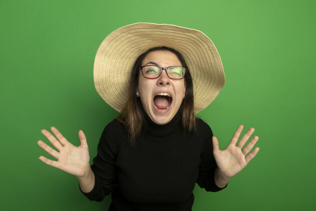 女孩年轻漂亮的女人戴着夏天的帽子 穿着黑色高领毛衣 戴着眼镜 高举双手站在绿色的墙上大声喊叫绿色手站着