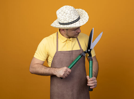 花园年轻的园丁穿着连体衣 戴着帽子 手持树篱修剪器 站在橙色的墙上看着它 很好奇制服人橙色