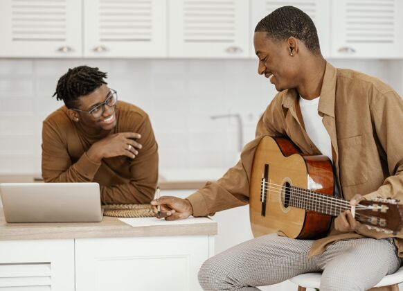 熟练程度笑脸男音乐家在家里的椅子上弹吉他和使用笔记本电脑人才男性音乐家
