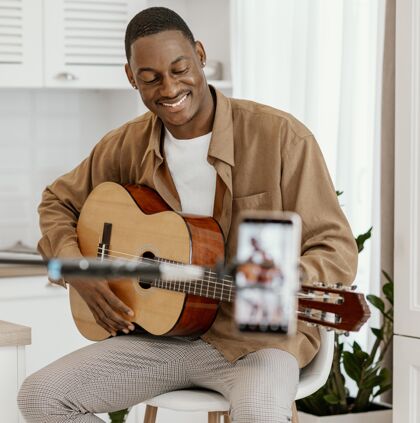 男性笑脸男音乐家在家里的椅子上弹吉他和录音与智能手机男人娱乐广场