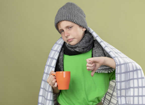 光生病的小男孩穿着绿色t恤 戴着暖和的围巾 帽子裹着毯子 手里拿着一杯热茶 大拇指朝下站在明亮的墙上疾病穿小