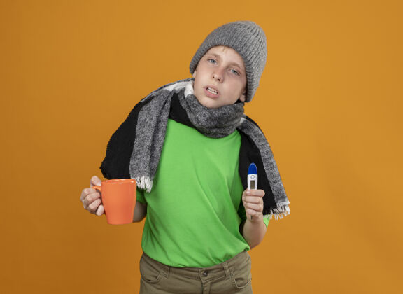 感觉生病的小男孩穿着绿色t恤 戴着暖和的围巾 戴着帽子 手里拿着一杯热茶和温度计 站在橙色的墙上感到不舒服 不舒服和不开心橘子不开心热