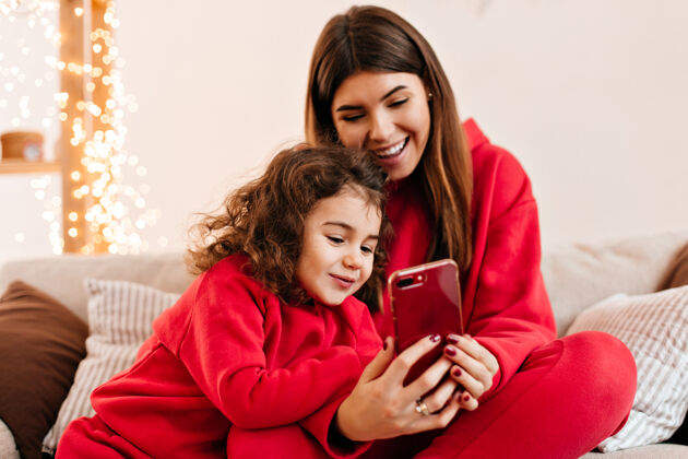 家庭无忧无虑的黑发女人带着女儿用智能手机一家人穿着红色衣服坐在沙发上的室内镜头童年微笑沙发