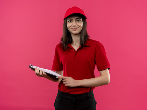马球穿着红色马球衫 戴着帽子的年轻送货女孩微笑着拿着剪贴簿站在粉红色的墙上交货微笑看