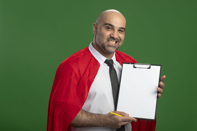 人穿红斗篷的超级英雄商人展示着空白页的剪贴板 站在绿色的墙上 面带微笑地看着前面页面超级斗篷