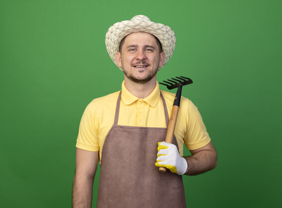 人年轻的园丁穿着连体衣 戴着帽子 戴着工作手套 拿着小耙子 面带微笑地站在绿色的墙上看着前面绿色穿站着