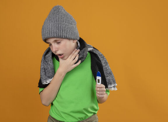 生病生病的小男孩穿着绿色t恤 戴着暖和的围巾和帽子 站在橙色的墙上 体温计显示不舒服 生病和不愉快的咳嗽男孩不开心秀