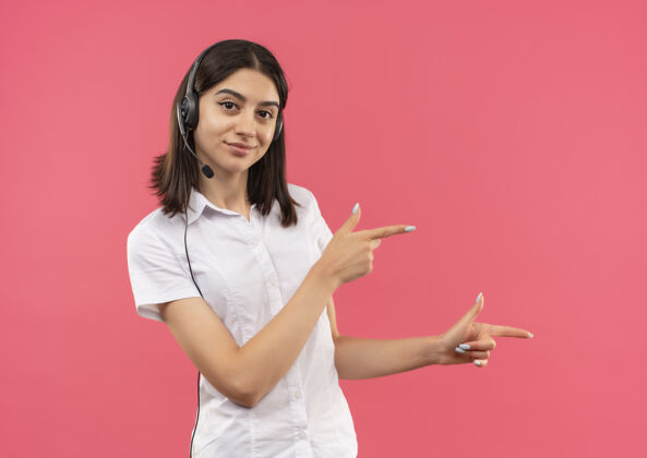 女孩一个穿着白衬衫戴着耳机的年轻女孩 手指着面带微笑地站在粉色的墙上人指向年轻人