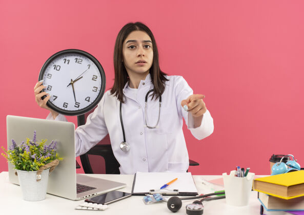 年轻年轻的女医生穿着白大褂 脖子上戴着听诊器 拿着挂钟 手指指向前面 坐在桌旁 笔记本电脑盖在粉红色的墙上 看上去很困惑手指墙壁拿着