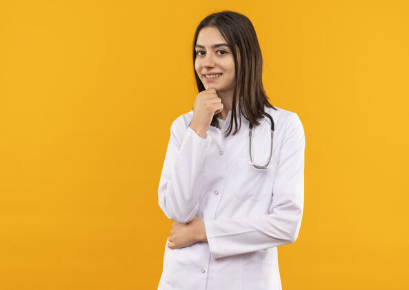 脖子年轻的女医生穿着白大褂 脖子上戴着听诊器 站在橙色的墙上 困惑地看着前方年轻人工作医院