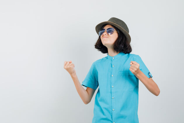 快乐身着蓝色衬衫 戴着帽子 戴着太阳镜 看起来很开心的年轻女士展示着赢家的姿态时尚欢呼室内
