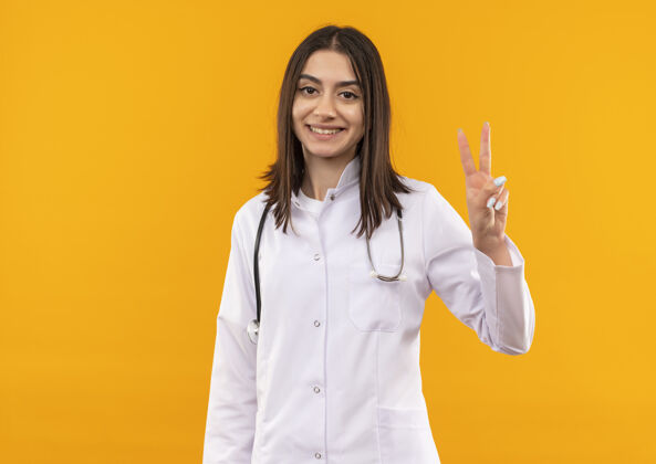微笑身穿白大褂 脖子上戴着听诊器的年轻女医生面带微笑 站在橙色的墙上 露出胜利的标志看女人表演