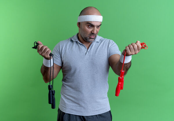 人戴着头巾的成熟的运动型男人拿着两条跳绳困惑地看着他们站在绿色的墙上有疑问姿势教练人