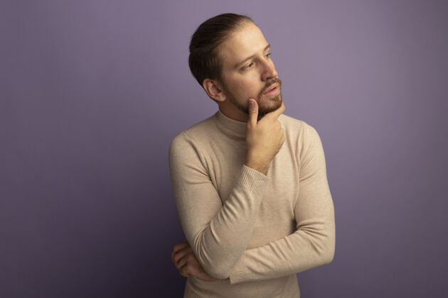 胡须穿着米色套头衫的年轻帅哥 手放在下巴上 站在淡紫色的墙上思考着思考丁香毛衣