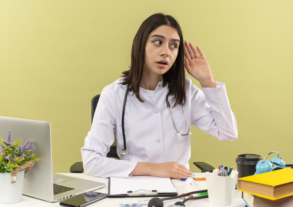 听诊器年轻的女医生穿着白大褂 脖子上戴着听诊器 手放在耳朵旁 一边看一边试图听坐在桌边的人说话 笔记本电脑放在浅色的墙上附近医生脖子