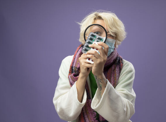 放大镜生病的不健康的女人 短头发 戴着暖和的围巾和面罩 站在紫色的墙上 透过放大镜看药片玻璃通过生病