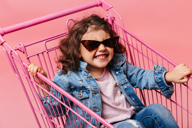微笑幸福的学龄前儿童坐在购物车里在粉色背景下 穿着牛仔裤的欢笑的孩子玩得很开心粉色隔离牛仔顾客
