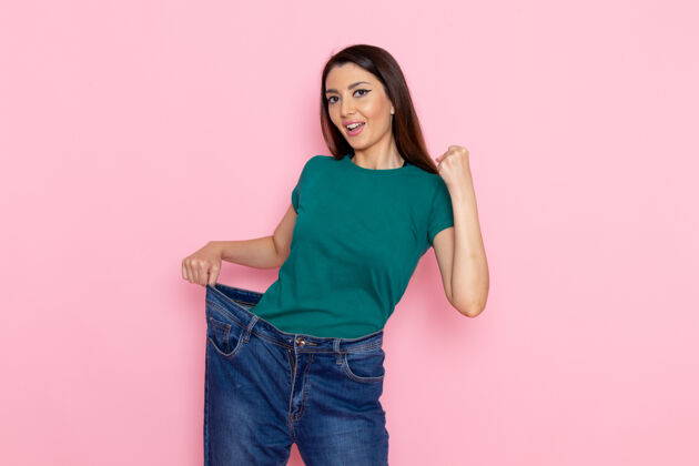 运动正面图身着绿色t恤的年轻女性在浅粉色墙壁上检查腰部腰部运动锻炼美丽苗条运动员女性腰部锻炼牛仔裤