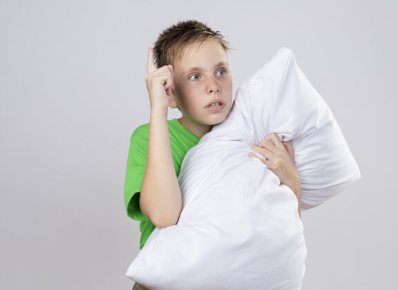 站生病的小男孩穿着绿色t恤 身体不适 抱着枕头 站在白色的墙上 焦急地看着一边旁边拥抱男孩