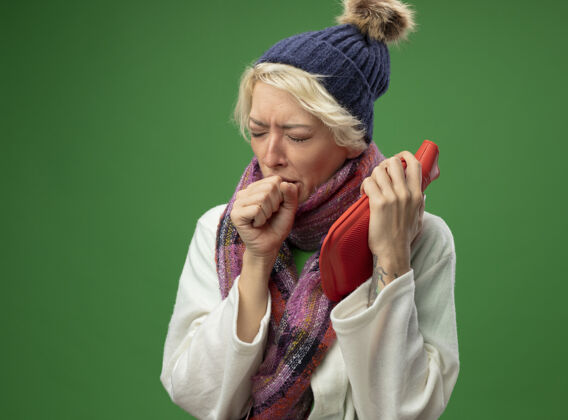 瓶子有病的不健康的妇女 短发 戴着暖和的围巾和帽子 感觉不舒服 拿着水瓶 站在绿色的墙上咳嗽 患流感立场温暖围巾