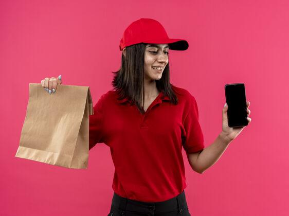 脸穿着红色马球衫 戴着帽子 拿着智能手机和纸包的年轻送货女孩站在粉色的墙上 面带微笑地看着一旁移动年轻微笑