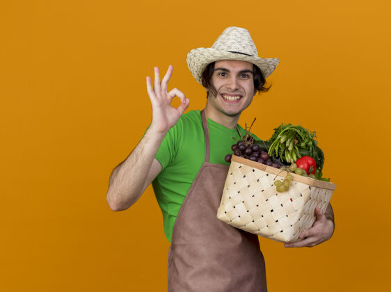 橘子年轻的园丁 围着围裙 戴着帽子 手里拿着装满蔬菜的箱子 满脸笑容 站在橘色的墙上 挂着“ok”的牌子帽子蔬菜花园