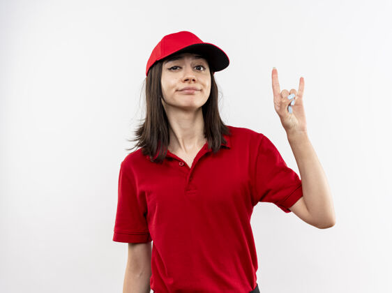 怀疑年轻的送货员身穿红色制服 戴着帽子 带着怀疑的微笑 站在白墙上的是岩石符号年轻穿显示