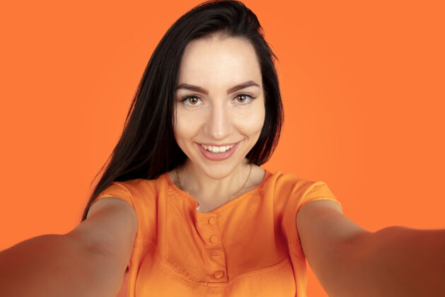 人橙色工作室背景上的白种人年轻女子肖像穿着衬衫的漂亮黑发女性模特人类情感的概念 面部表情 销售 广告空间自拍和微笑面部财务女商人