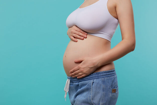 健康年轻孕妇穿着白色运动上衣和牛仔裤 双手高举在腹部 在蓝色背景上摆姿势的不规则照片自然温柔站