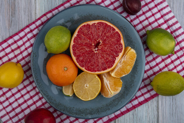 酸橙顶视图：半个葡萄柚 橘子和削皮的楔子放在盘子里 柠檬 酸橙和桃子放在红色方格毛巾上桃子橘子盘子