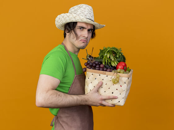 抱着年轻的园丁 围着围裙 戴着帽子 手里拿着装满蔬菜的箱子 站在橘色的墙上皱着眉头 看起来很不高兴看蔬菜橘子