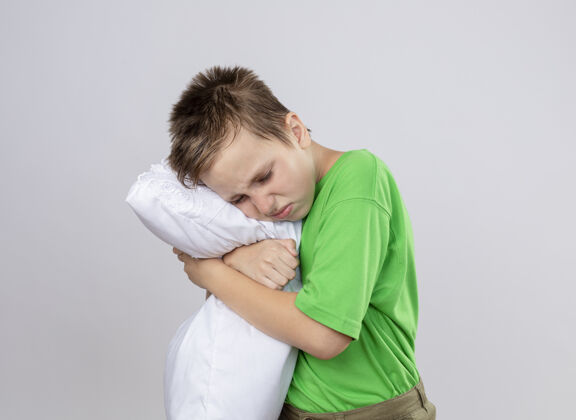 枕头生病的小男孩穿着绿色t恤 感觉不舒服 抱着枕头站在白墙上受冻疾病不适感觉