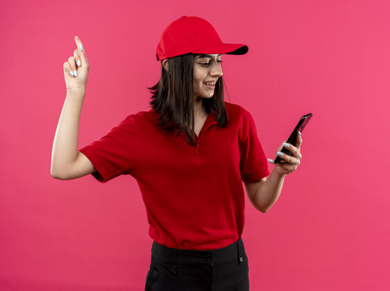 站穿着红色马球衫和帽子的年轻送货女孩看着她的智能手机屏幕 手指露出自信的表情 脸上带着微笑站在粉色的墙上穿微笑脸