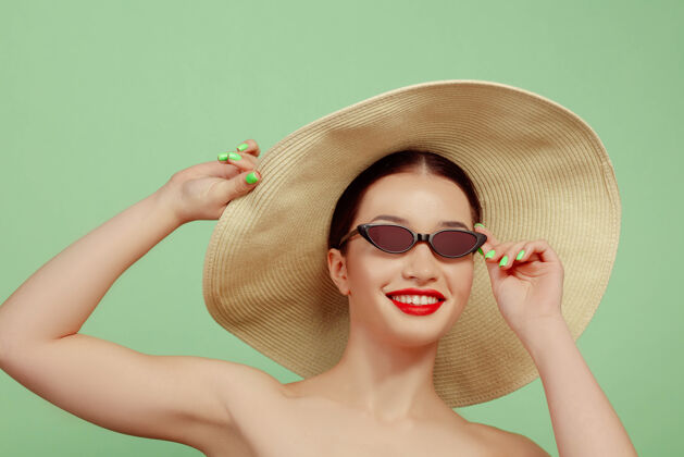 帅气美丽女人的肖像 明亮的妆容 帽子和太阳镜在绿色的工作室背景上时髦和时尚的制作和发型夏天的颜色美丽 时尚和广告概念微笑自信保持人