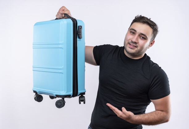 假期身穿黑色t恤的年轻旅行家站在白色的墙上 带着自信的微笑展示手提箱信心姿势旅行